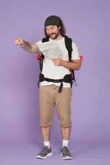 Man tourist in casual with backpack holding paper map, pointing with smiling face. Isolated on purple background. Portrait of male traveler.