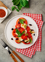 Italian caprese salad of tomatoes, mozzarella and basil on a white ceramic plate, red and white background, a bowl with red spices nearby, close-up, top view