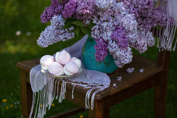 A bouquet of lilac flowers in a vase and a saucer with marshmallows on a chair in the garden against a background of greenery, close-up, soft focus. Beautiful spring card.