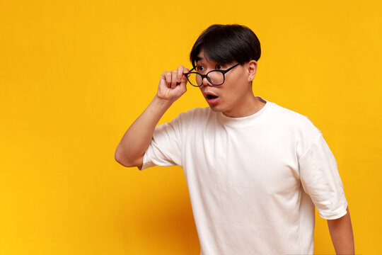 young shocked asian guy in glasses looks surprised at copy space on yellow isolated background