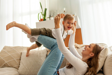 young mother holds and lifts her little son on the sofa, woman plays with her child at home, 2 year...