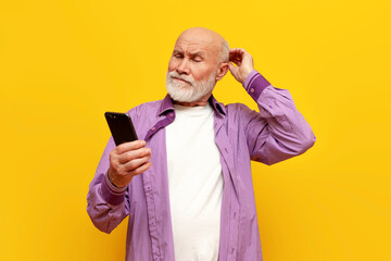 puzzled old bald grandfather in purple shirt uses smartphone on yellow isolated background, confused old pensioner - obrazy, fototapety, plakaty