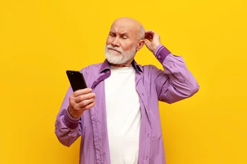 Fotobehang puzzled old bald grandfather in purple shirt uses smartphone on yellow isolated background, confused old pensioner © Богдан Маліцький