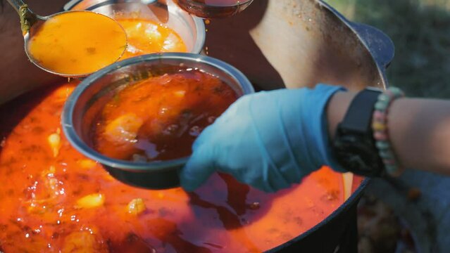 The cook ladles red meat soup into metal bowls. Close-up top view of huge cauldron of bograch or borscht outdoors and female hands holding kitchen ladle. Camping season