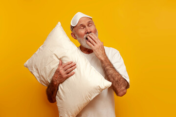 sleepy old bald grandfather in pajamas and sleep mask hugs pillow and yawns on yellow isolated background
