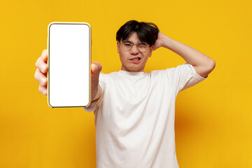 puzzled young guy asian student shows blank smartphone screen on yellow isolated background and...