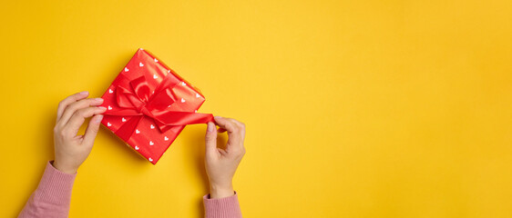 Two female hands hold a red box tied with a ribbon on a yellow background, top view.