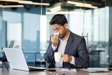 Young Hindu businessman sneezes and has a runny nose while working in the office at the computer,...