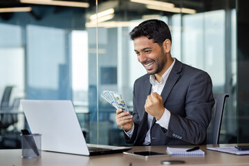 Happy Indian man sitting at a desk in the office in front of a laptop with a wad of money in his...