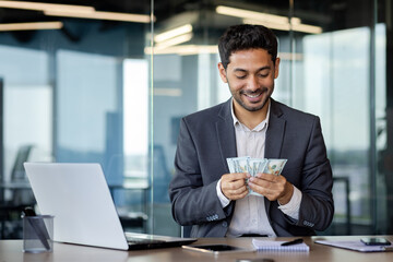 Happy Arab worker in office sitting in front of laptop contentedly counting dollars earned online,...