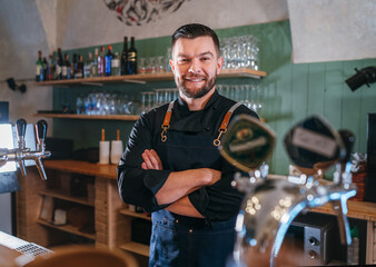 Portrait of happy smiling bearded barman dressed in a black uniform with an apron at bar counter...