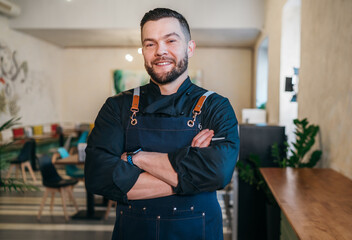 Portrait of happy smiling small business owner dressed in a black chef uniform with an apron in his cozy restaurant hall. Successful people, hard work, consumer cafes and restaurants industry concept. - obrazy, fototapety, plakaty