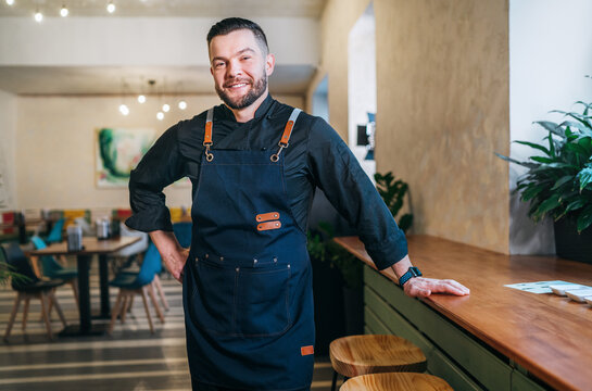 Portrait of happy smiling small business owner dressed in a black chef uniform with an apron in his cozy restaurant hall. Successful people, hard work, consumer cafes and restaurants industry concept.