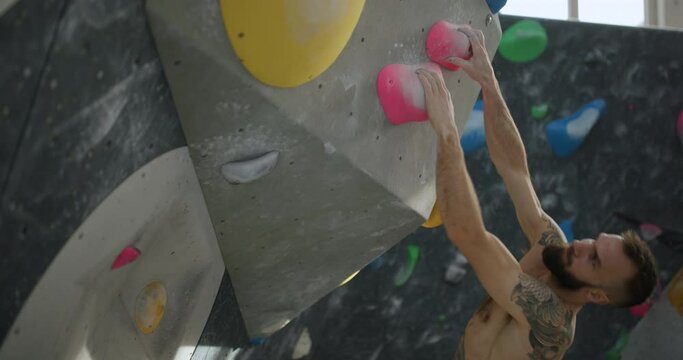 Fit young man with no shirt and tattoos using handholds to climb up an artificial rock wall in a bouldering gym
