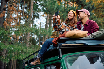 Obraz premium Happy couple using mobile phone and taking pictures while sitting on top of their camper trailer in woods.