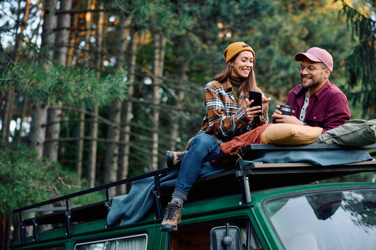 Happy couple using cell phone while sitting on top of their van during camping in woods.