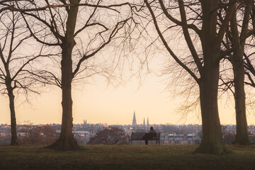 Moment of solitude as a distant figure sits on a quiet park bench during a cold winter sunset or sunrise at Inverleith Park in Edinburgh, Scotland, UK.