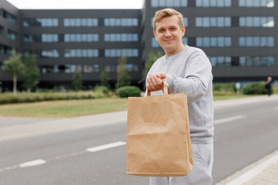 Man holding restaurant takeaway meal, Office food delivery service courier giving order package, standing near the office building. Mock up, craft paper bag