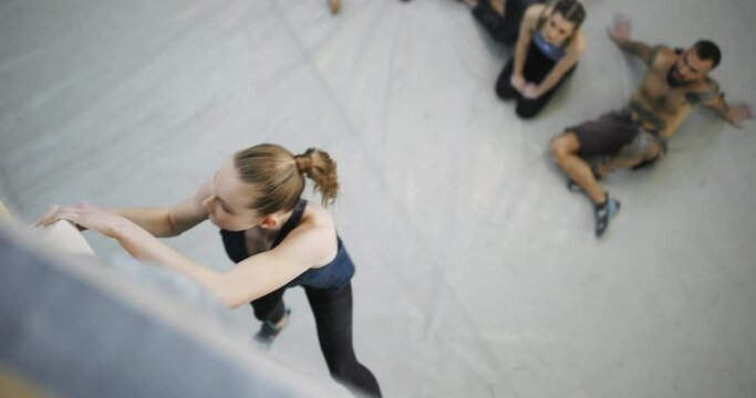 Fit young woman with chalked hands climbing an artificial rock wall in a gym with friends watching from the ground