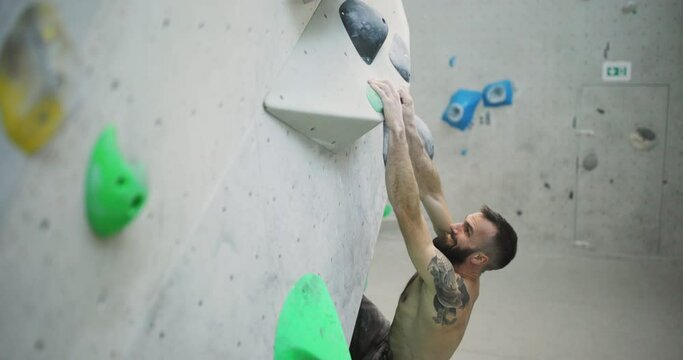 Fit man with tattoos using hand and footholds to climb up an artificial wall in a bouldering gym