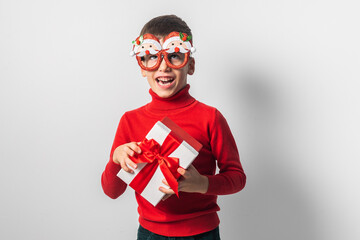 Funny grimacing child in Santa red sweater holding Christmas gift in hand. Christmas festive holidays concept.