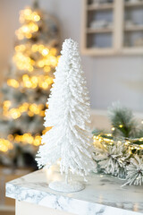 White artificial Christmas tree on a table on background of garland lights. Vintage New Year decorations. Stylish interior in light colors. Christmas mood. Celebrating of New Year