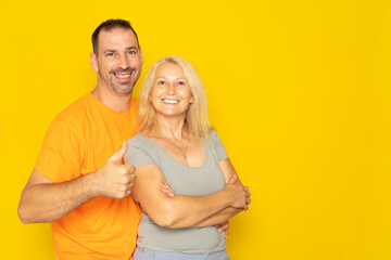 Happy adorable Caucasian couple in their 40s hugging and raising thumbs up in sign of approval and trust, isolated on yellow studio background.