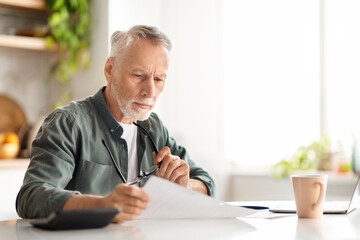 Elderly man reviewing documents while sitting at kitchen desk - obrazy, fototapety, plakaty