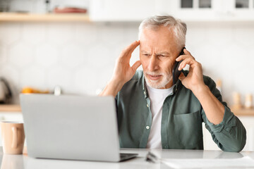 Stressed senior man using phone and laptop, sitting at desk in kitchen