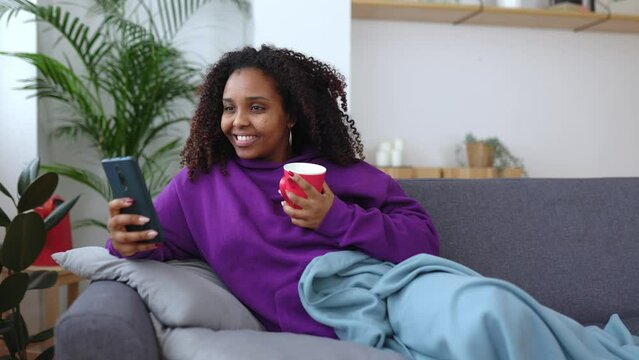 Young african american woman relaxing on sofa using mobile phone in the morning. Happy black female drinking coffee while enjoying holiday morning winter vacation resting on couch chatting on cell.