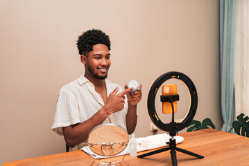 Young latin man creating skincare content with smartphone and ring light