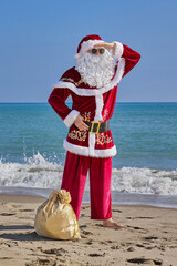Santa Claus at sea on a hot summer day. Santa Claus stands barefoot on a sandy beach in front of the sea. Bag of gifts on the sand.