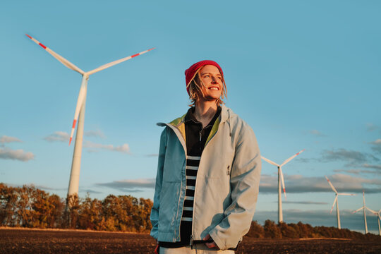 Smiling alternative woman looking away in denim jacket by wind turbines