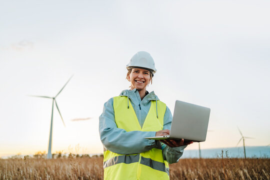 Engineer looking at camera with laptop at wind farm during sunset