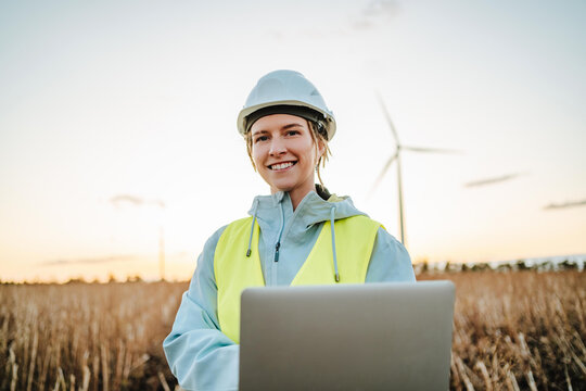 Smiling engineer with laptop near wind turbines at dusk