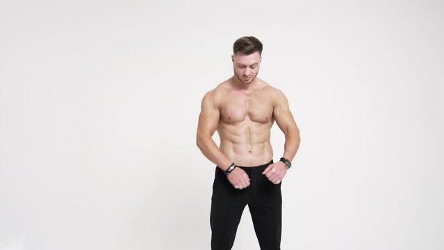 Sportive man with naked torso holding hands in pockets of black trousers. Man crosses hands on chest smiling to camera. White backdrop.