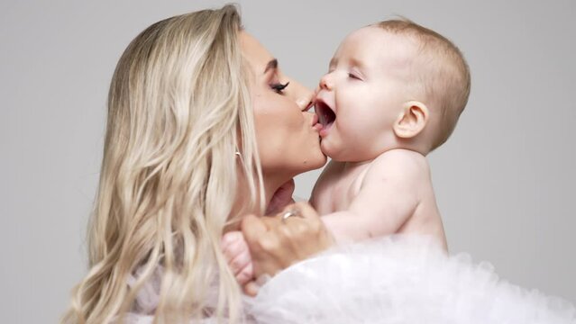 Loving mom kisses her adorable little baby. Blonde woman holds her naked infant son in studio. White backdrop.