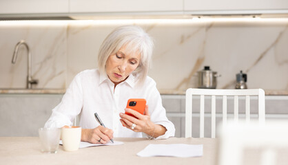 Elderly woman calculates finances and fills out paperwork using internet and smartphone