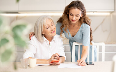 Positive young woman and her senior mother sitting at table in kitchen, doing family financial paperwork