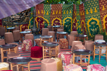 Arabian tea room with small tables and coloured stools, silver trays and in the background a tapestry full of arabesque vegetal and geometrical motifs in many colours. - obrazy, fototapety, plakaty