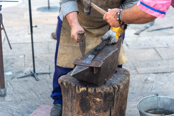 Detail of the hands of two blacksmiths in brown leather aprons and gloves forging a piece with a hammer on an iron anvil in the street at a street market.