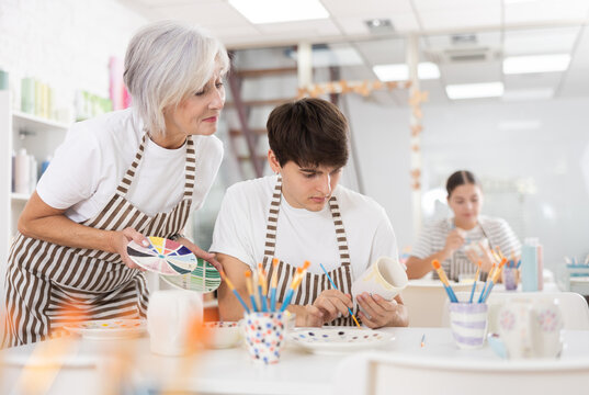 Interested Elderly Woman Assisting Young Adult Son Working On Painting Ceramic Mugs In Family Workshop. Concept Of Intergenerational Exchange Of Pottery Craftsmanshi