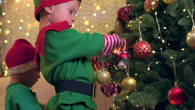 two children brothers boys in elf costume decorate Christmas tree together. happy toddlers celebrate Christmas and New Year. kids in red Santa hats hang decoration balls Christmas tree together
