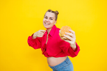 A cute pregnant woman in a pink shirt is eating a fast food burger. A pregnant woman on a yellow background with an appetizing burger in her hands. Harmful food for pregnant women.