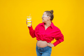 A cute pregnant woman in a pink shirt drinks water from a plastic cup. A pregnant woman on a yellow background with a glass in her hands. Harmful water for pregnant women.