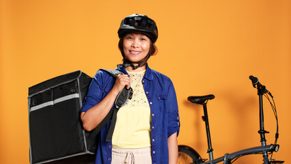 Portrait of happy smiling asian bike rider holding thermal backpack, isolated over orange studio background. Friendly courier express delivering takeaway food to customer