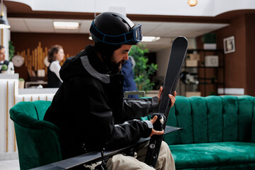 Seated caucasian customer in hotel reception adjusts ski gear, wearing winter attire. Ready for skiing excursion, male traveler inspects wintersports equipment in preparation of snowy slopes adventure