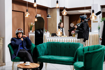 Asian female guest seated on sofa securing snow helmet while young man with skiing skis doing...