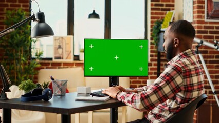 African american teleworking freelancer inputing data on chroma key mock up computer green screen in apartment living room. Relaxed remote employee performing tasks while working from home