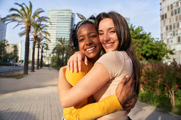 Portrait multi-ethnic girlfriend young couple posing hugging for photo outdoor in summer. Happy Caucasian woman and smiling African girl embracing on city street. Cheerful females in loving attitude.